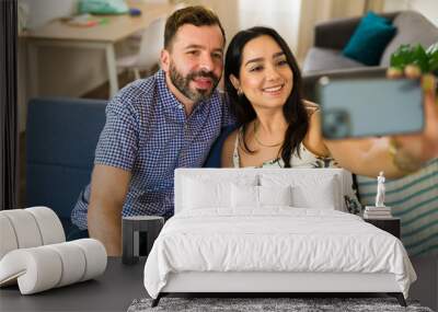 Happy young couple taking a selfie on their sofa at home, showing off their love and bond Wall mural