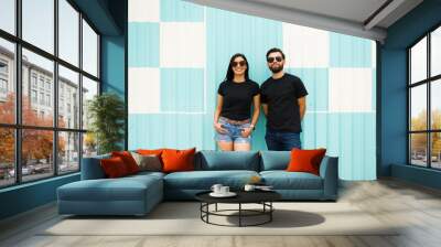 Latin couple in sunglasses, posing confidently with space for logo on their black t-shirts against a turquoise and white striped backdrop Wall mural