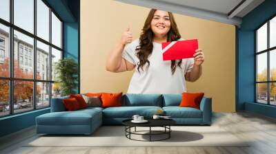 Pretty Hispanic plus-size woman in a photo studio holding a red gift card and giving a thumbs up sign Wall mural