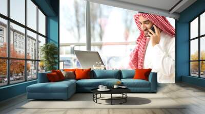 Happy arab man using a laptop and talking on phone in a bar Wall mural