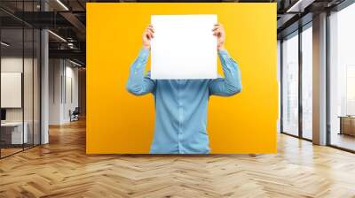 a person standing against a bright yellow background, holding a blank white poster in front of their face Wall mural