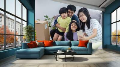 Asian family with two little kids sitting on couch at home together, taking selfie, smiling at smartphone camera, mom holding phone. Medium shot. Family photo or communication concept Wall mural