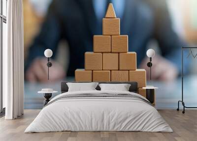 A pyramid of wooden blocks stands in front of a businessman, symbolizing structure, stability, and growth Wall mural