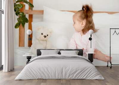 A baby girl in a pink bodysuit on the bed in the bedroom smiling or laughing, a cute little child at home with a teddy bear, a place for text Wall mural