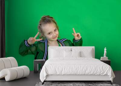 a cheerful girl with curly hair looks at the camera and shows two fingers, standing on a green background. space for text Wall mural