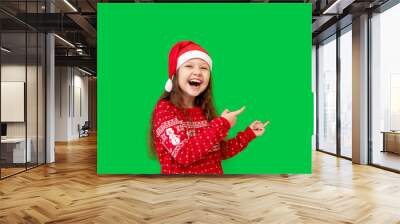 a happy little girl child in winter clothes, a Santa Claus hat and a red sweater on a green isolated background shows her fingers up at something and smiles, a place or space for text Wall mural