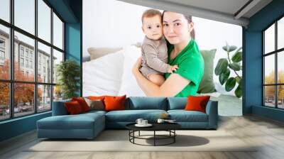 a young mother in green clothes holds her baby boy, hugs and kisses on the bed in a bright bedroom, motherhood Wall mural