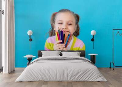 portrait of a cute little girl with colored pencils in a striped jacket. The concept of education and drawing. Photo studio, blue background, place for text Wall mural