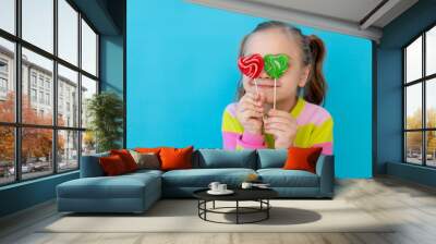 portrait of a cute little girl with lollipops in a striped jacket covering her eyes with them, a large candy on a stick. The concept of sweets and confectionery. Blue background, space for text Wall mural