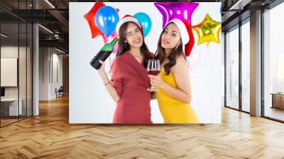 Two young happy Asian women celebrating the Christmas and new year 2022 and drinking red wine. Two young girls in red and yellow dress sip wine in a white scene with multi colored balloons. Wall mural