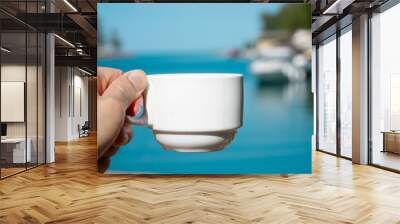 Man holds a white coffee cup in his hand on the background of the sea and boat. Wall mural