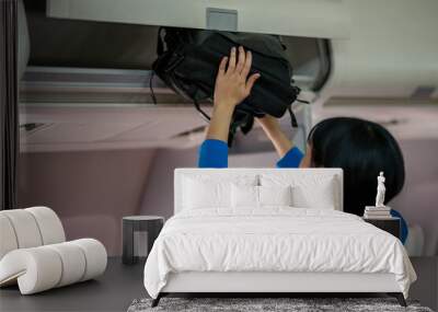 A traveler in a blue jacket is placing her carry-on bag into the overhead storage compartment on an airplane. Wall mural