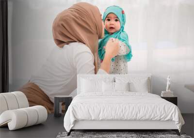 Happy muslim mother and adorable little baby daughter in hijab on bed in white bedroom at home. Muslim mom hands holding baby for practice standing on bed at indoors. Love of mother and baby concept. Wall mural