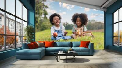 Two African dark skinned children boy and girl sitting on field grass and eating fruits together in parks and outdoors. Wall mural