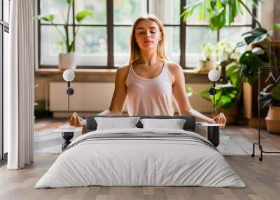 A woman practices meditation in a serene living room surrounded by plants, representing a peaceful and healthy lifestyle. Wall mural