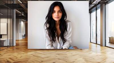 Portrait of a young woman with long brunette hair sitting indoors against a white wall, wearing a casual white shirt and jeans. Wall mural