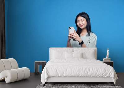 Young beautiful woman typing a text message or scrolling a social network using a smartphone and smiling holding a phone in her hands isolated on a blue background Wall mural