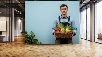 Calm young Caucasian farmer in work clothes holding a basket of vegetables against a light blue background Wall mural