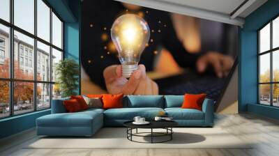 Man in black holding a bright orange light bulb with a laptop, indicating answering questions or thinking of new ideas or creativity, as well as business startups. Wall mural