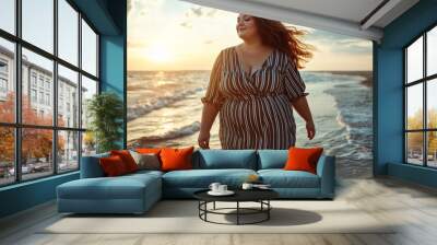 Plus-size woman enjoying a peaceful sunset walk along the beach, with waves gently touching her feet and a serene sky in the background. Wall mural