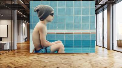 A young boy wearing a gray beanie and sunglasses, sitting quietly by the edge of a pool with a blue tiled background, reflecting a calm and thoughtful mood Wall mural