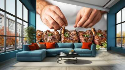 Close-up of a chef's hands arranging grilled lamb chops with rosemary sprigs on a white platter. Wall mural