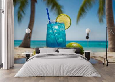 A refreshing image of a glass of blue cocktail, placed on a sandy beach with a clear blue sky and ocean in the background and with slices of lemon and fresh mint leaves. Wall mural