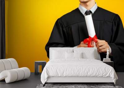 A man wearing a graduation gown holds a diploma in his hands on a yellow background. Wall mural