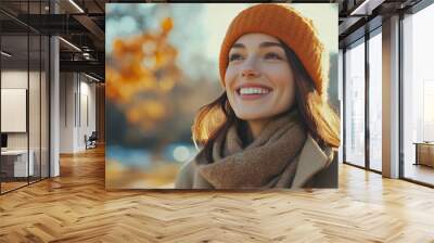 A young woman wearing a warm orange hat and scarf smiles brightly at the camera, enjoying the autumn weather. Wall mural