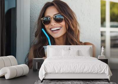 A young woman with long brown hair, wearing sunglasses, smiles broadly as she enjoys a cold beverage on a summer day. Wall mural