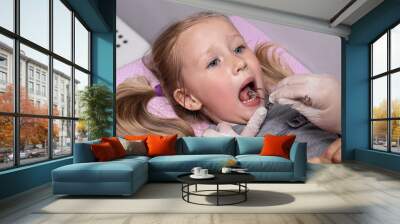 a small blonde girl of 4 years old at a speech therapist's reception, working with the muscles of the cheeks and tongue with special tools Wall mural