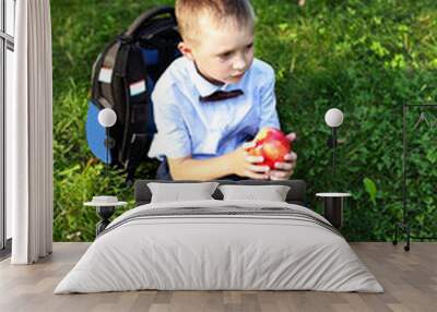 charming blond boy 6 years old in a light shirt and bow tie with a backpack on his back sits on the green grass in the Park and eats an Apple Wall mural