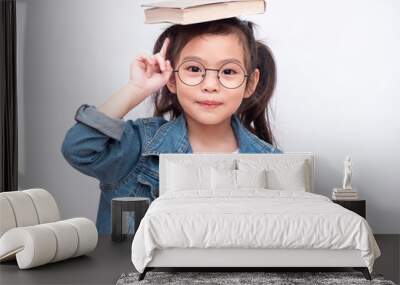 Asian little cute girl wearing glasses and put the book on head. Preschool lovely kid with the book on her head and pointing finger up. Learning and education of kid. Wall mural