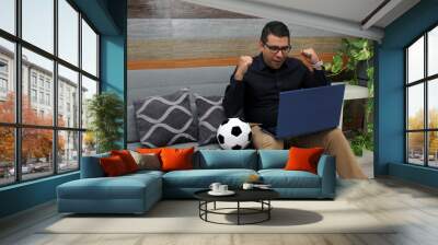 Latino adult office man watches football games on his work laptop during office hours in the morning, he sees him excited, nervous, surprised next to his soccer ball Wall mural