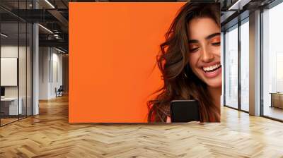 Photo of an elegant young woman in her 20s with long wavy hair, happy smiling while using a mobile phone on an orange and black background with copy space. Wall mural