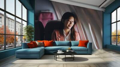 Portrait of a successful Asian businesswoman or entrepreneur in a formal suit on an airplane sitting in business class using a phone, computer laptop. Travel in style, work with grace. Wall mural