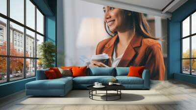 Successful businesswoman smiling holding coffee cup at office. Confident business woman standing drinking coffee in the office. Woman standing by the window. Wall mural