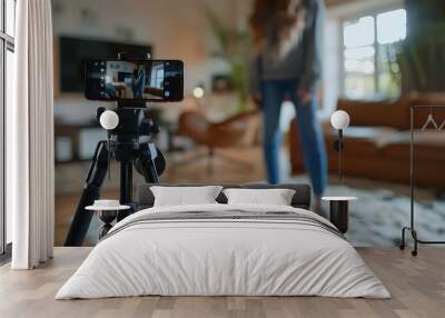 A young woman films a video on her smartphone while standing in a living room with a couch and a window in the background. Wall mural