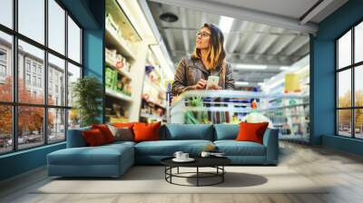 Groceries, phone and woman in supermarket or grocery store using app for shopping choice of food on a shelf. Website, retail and young customer with a decision by a refrigerator with a smartphone Wall mural