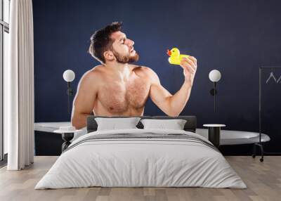 Handsome naked muscular adult happy man taking bath and playing with toy ducks, on dark-blue background Wall mural