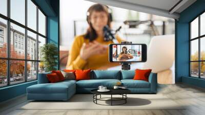 Female vlogger live streaming podcast using microphone, laptop and cellphone on tripod. Selective focus on smartphone camera screen with woman podcaster recording and broadcasting live video. Close-up Wall mural