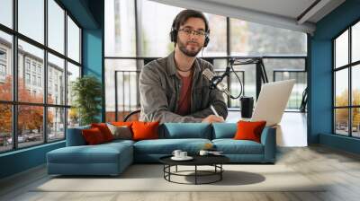 Portrait of cheerful young man host recording podcast in studio. Handsome smiling guy wearing headphones and glasses sitting at table with laptop and microphone, looking at camera, using pen, notebook Wall mural