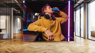 Portrait of young professional cyber sportsman sitting at gaming desk at home and drinking caffeine energy drink to concentrate on online video shooter game at night. Neon coloured room. Cyber sport Wall mural