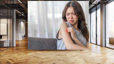 Asian woman with a disgusted expression touching her arm, feeling discomfort or itchiness on a sofa, health and skin irritation issue, unpleasant feeling, young female showing reaction of sensitivity Wall mural