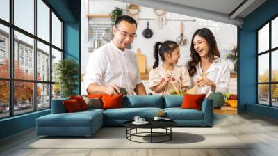 Portrait of enjoy happy love asian family father and mother with little asian girl daughter child having fun help cooking food healthy eat together with fresh vegetable salad ingredient in kitchen Wall mural