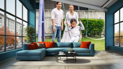 Portrait of enjoy happy love asian family father and mother with little asian girl smiling playing and pushing daughter on the swing moments good time at playground Wall mural