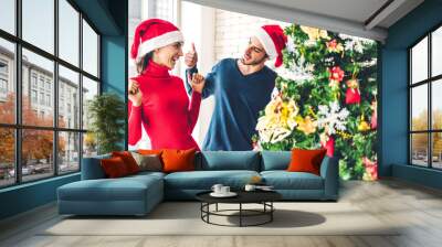 Romantic sweet couple in santa hats having fun decorating christmas tree and smiling while celebrating new year eve and enjoying spending time together in christmas time at home Wall mural