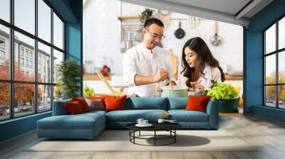 Young asian family couple having fun standing near stove and cooking together.Happy couple looking and smelling tasting fresh delicious from soup in a pot with steam at white interior kitchen Wall mural