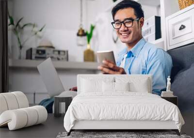 Young smiling asian man relaxing using laptop computer working and video conference meeting at home.Young creative man looking at screen typing message with smartphone.work from home concept Wall mural