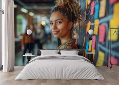 young woman with curly hair, wearing casual attire and smiling at the camera while standing in front of an office wall covered in colorful sticky notes modern open space office environment Wall mural